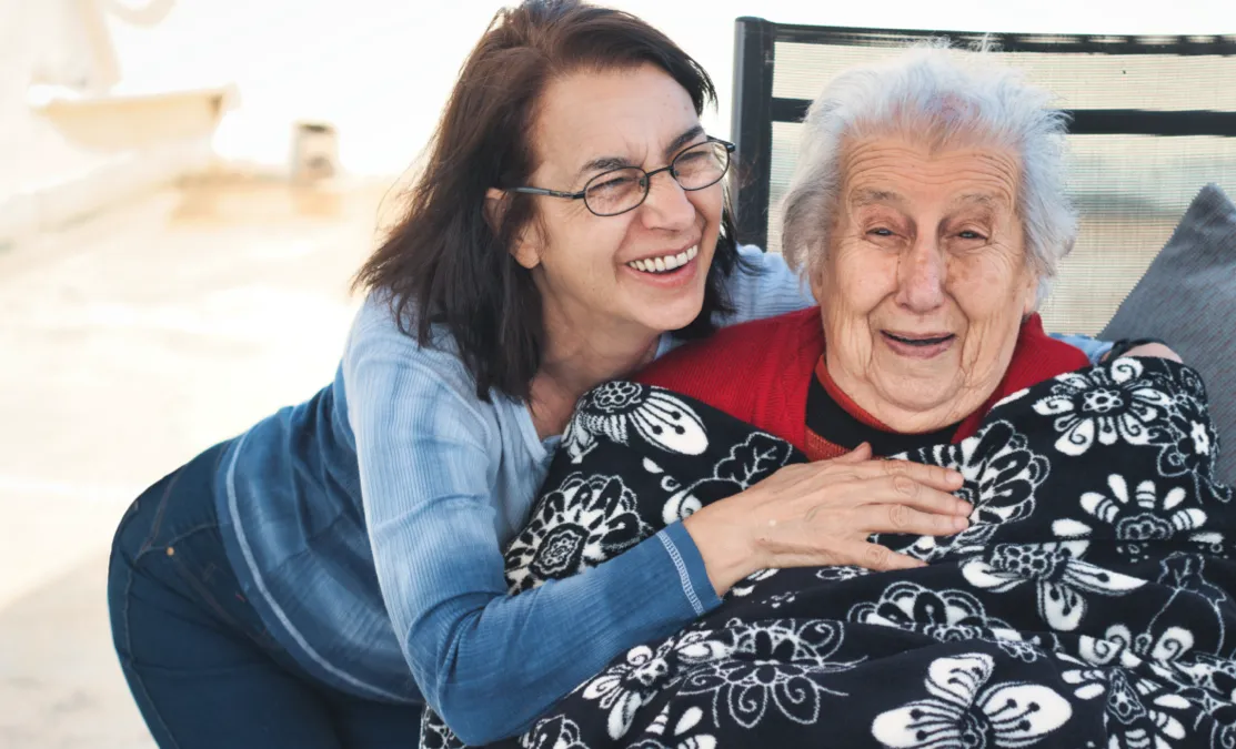 A joyful elderly woman with white hair, wrapped in a black-and-white patterned blanket, is sitting outside on a chair. Next to her is a younger woman wearing glasses and a blue shirt, leaning in close, laughing, and affectionately placing her hand on the elderly woman's shoulder. Both women are smiling warmly, sharing a happy moment together. The scene suggests a close bond between them.