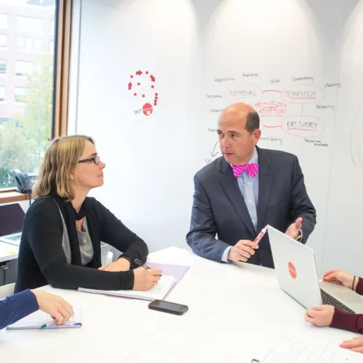 A group of professionals is engaged in a meeting around a whiteboard. A man in a suit and bright pink bow tie appears to be explaining something while holding a marker, gesturing towards a colleague. The woman across from him is listening attentively with a notepad in front of her. A laptop is open on the table, and some notes and diagrams are visible on the whiteboard in the background. The atmosphere suggests a collaborative work discussion or strategy planning session.