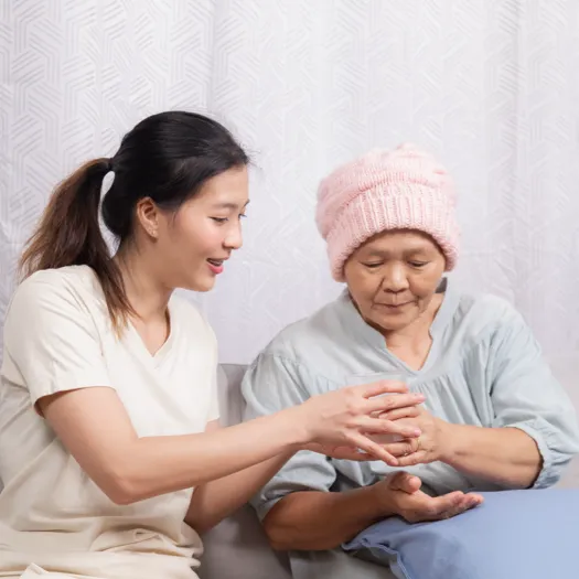 A young caregiver is gently assisting an elderly woman who is wearing a soft pink knit cap. The caregiver is smiling and holding the older woman's hands, offering support or guidance in a warm and caring manner. The elderly woman looks down at her hands, appearing thoughtful or focused. The setting suggests a calm and compassionate moment, possibly in a home or healthcare environment, highlighting caregiving or patient support.